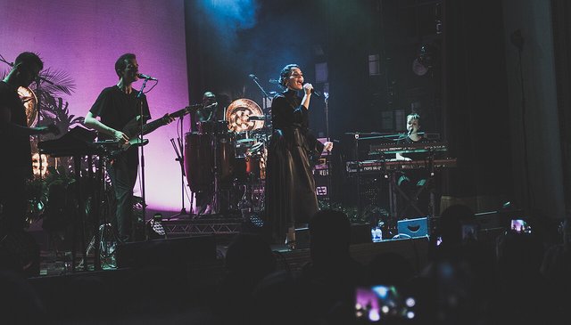 Jessie Ware singing into a microphone on a stage with a band, lit in purple and teal