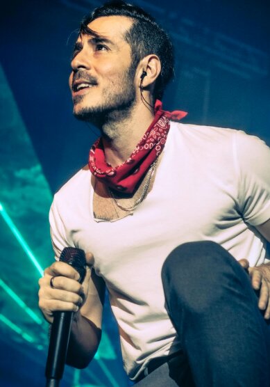 Singer in a white t-shirt and red bandana holding a microphone on a stage lit in blue and green