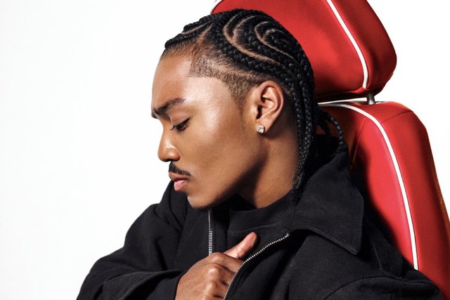 Profile portrait of a man with braided hair in a black jacket, seated against a red chair on a white background