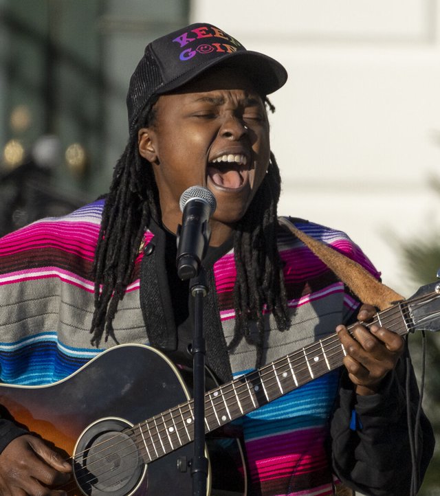 Joy Oladokun singing into a microphone while playing acoustic guitar in a striped serape