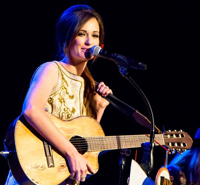 Kacey Musgraves singing into a microphone on stage under warm stage lights