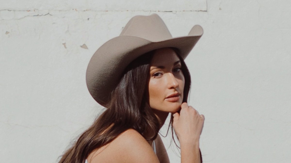 Kacey Musgraves in a tan cowboy hat, resting her chin on her hand against a white wall