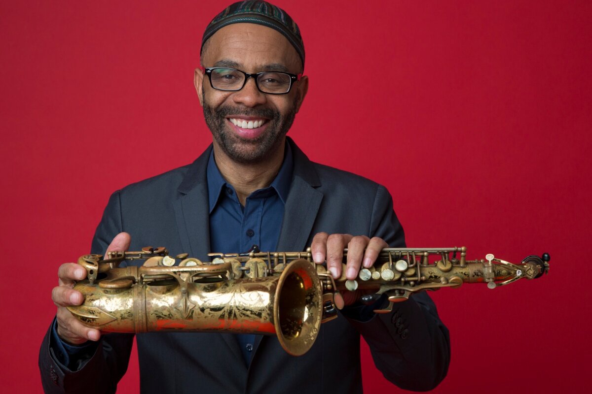 Kenny Garrett smiling in a kufi cap and glasses, holding his alto saxophone against a red backdrop