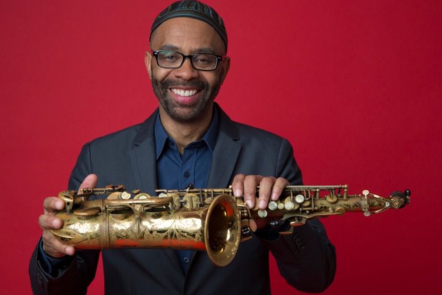 Kenny Garrett smiling in a kufi cap and glasses, holding his alto saxophone against a red backdrop
