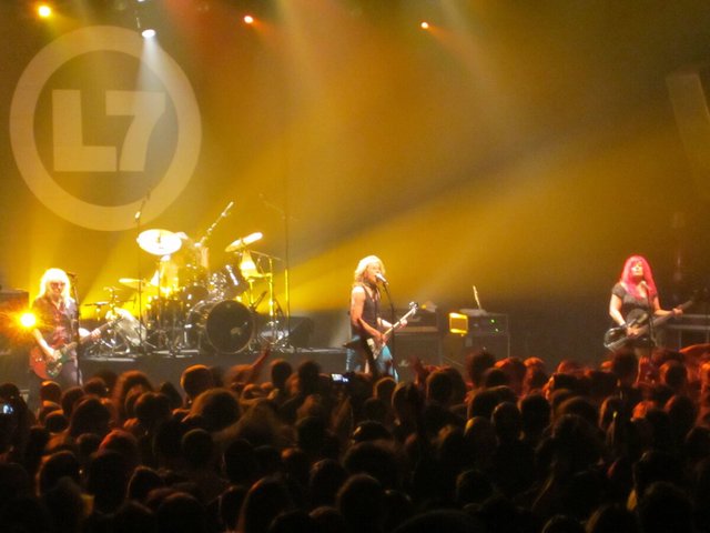 L7 performing on stage under yellow lights with the band's logo behind them and a crowd in the foreground