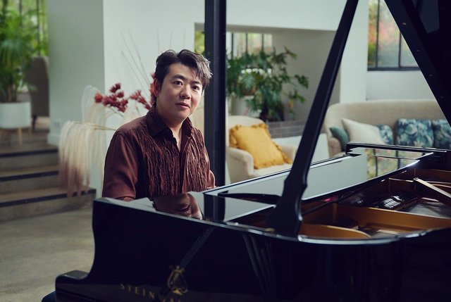Pianist Lang Lang in a brown shirt seated at a black Steinway grand piano with its lid open, in a bright room full of plants