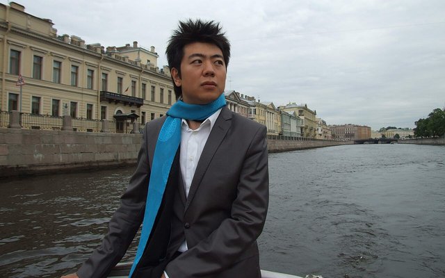 Pianist Lang Lang in a gray suit and blue scarf, seated on a boat on a city canal lined with pale historic buildings