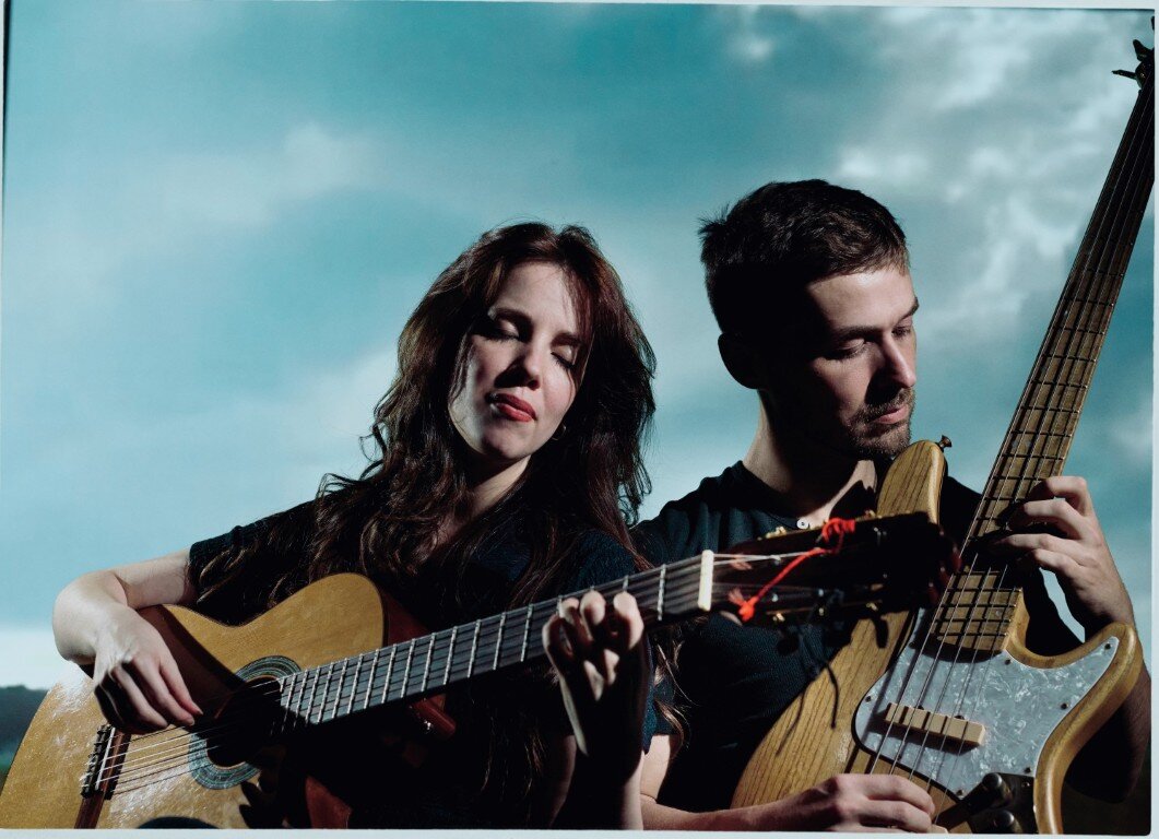 Lau Noah on classical guitar and Adam Neely on electric bass against a cloudy blue sky