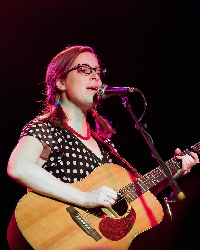 Laura Veirs singing into a microphone and playing an acoustic guitar on a dark stage