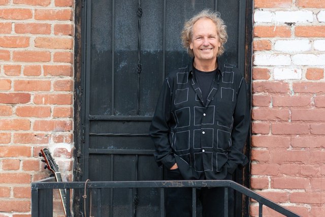 Lee Ritenour smiling in a black shirt beside a black door and brick wall, with an electric guitar at lower left