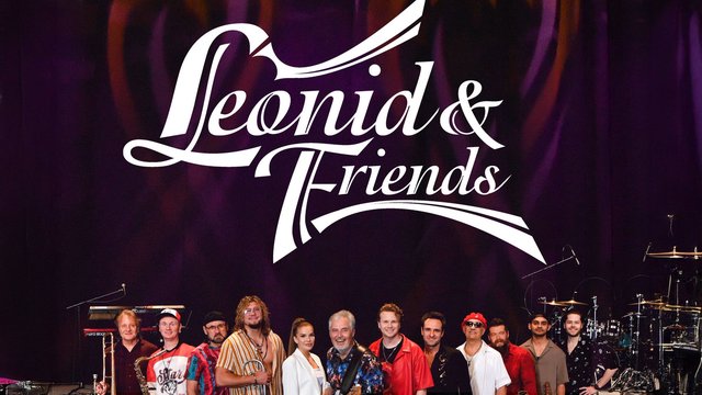 Leonid & Friends band members lined up on a stage with horns, keyboards and drums beneath the band's white script logo on a purple backdrop