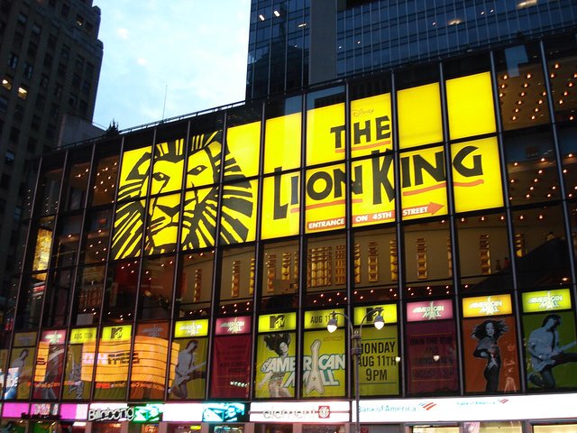 Glowing yellow Lion King billboard with the show's black lion face logo on a glass theater facade at dusk