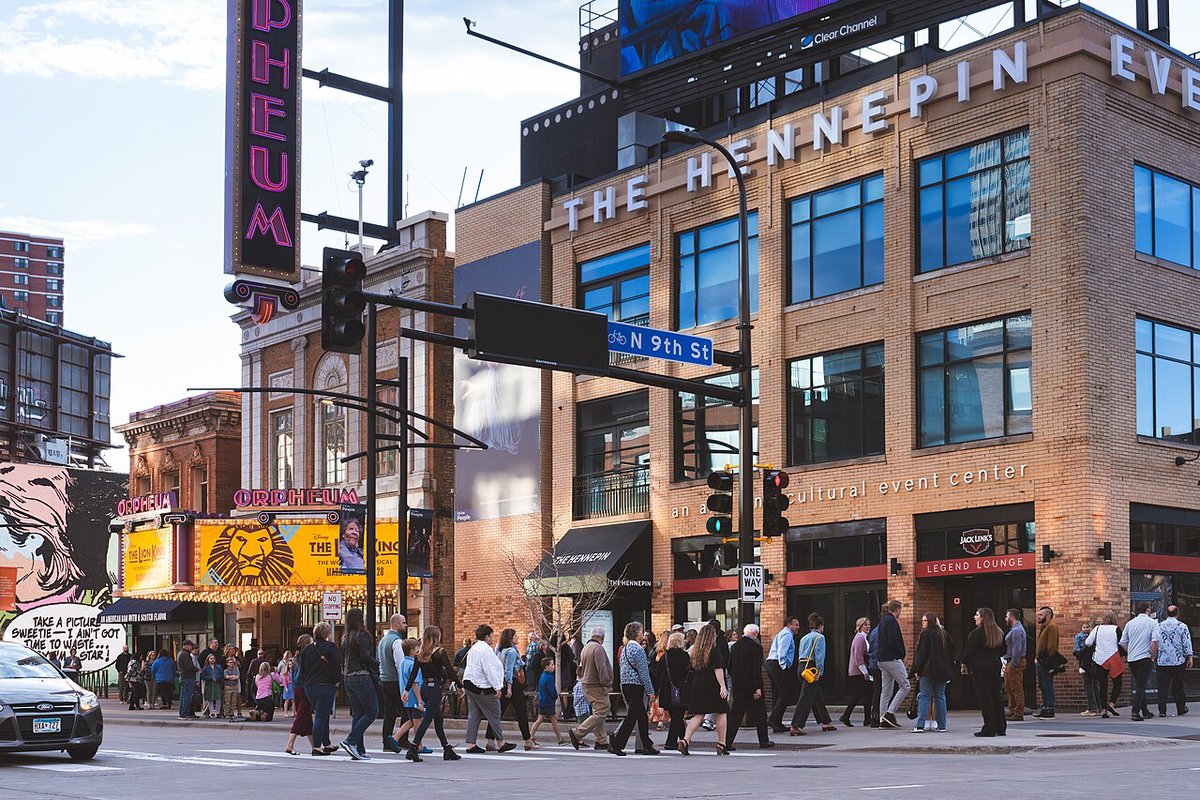 Theatergoers crossing the street past a marquee for the touring Lion King musical, with the gold lion logo lit above the doors