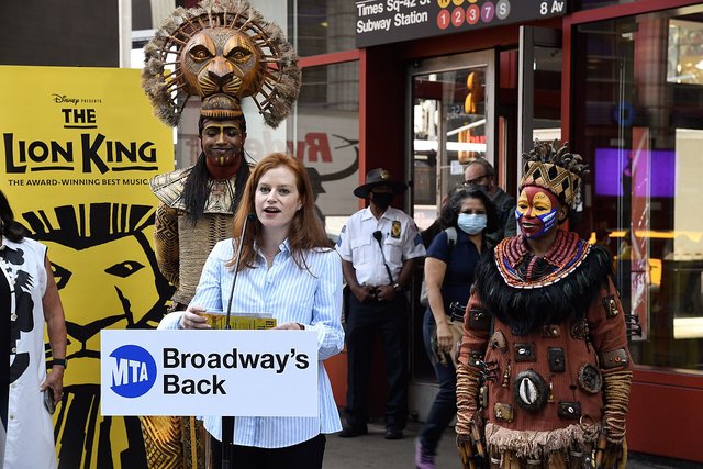 Lion King cast members in Mufasa's lion mask and Rafiki's beaded costume beside a yellow Lion King poster