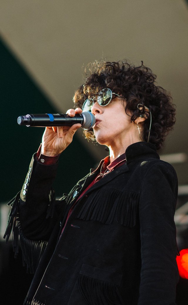 LP singing into a microphone on an outdoor stage, wearing round sunglasses and a fringed black jacket