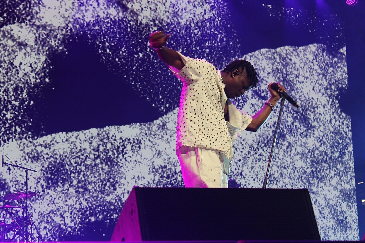 Lucky Daye in a studded white outfit leaning into a microphone on stage in front of a large video screen
