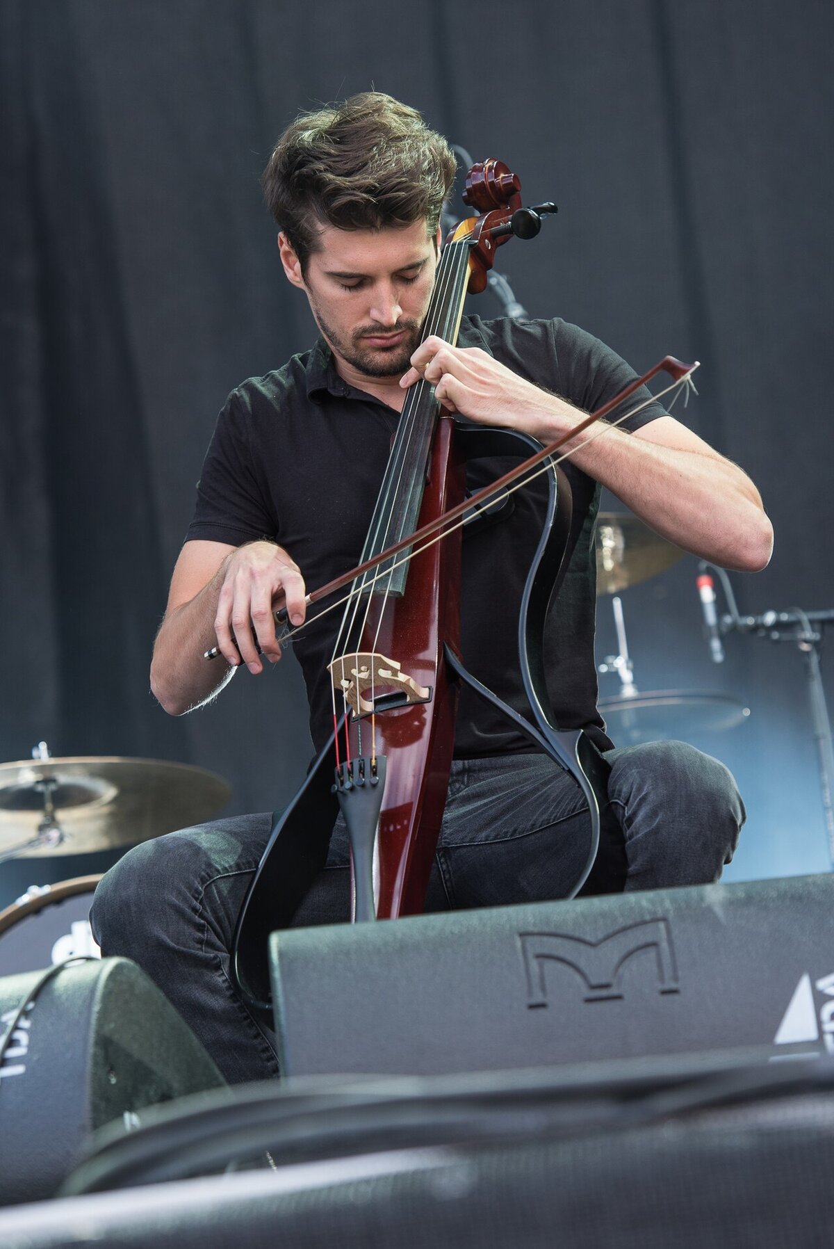 Cellist Luka Sulic playing an electric cello on stage with a drum kit behind him