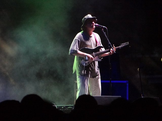 Mac DeMarco in a bucket hat playing electric guitar at a microphone on a smoky stage, with audience heads in silhouette