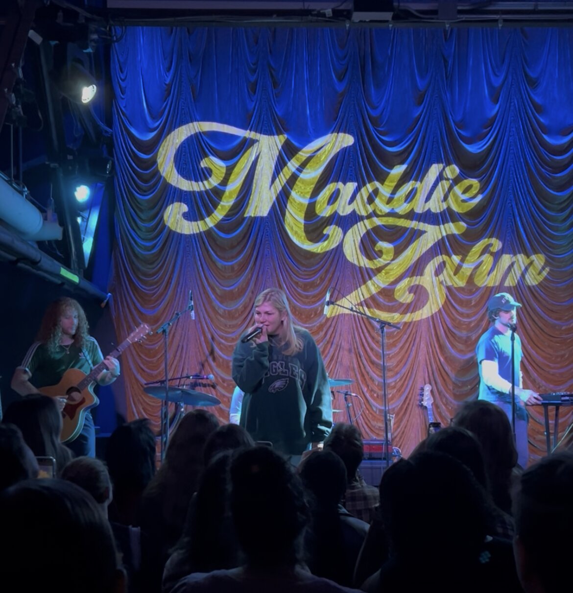 Singer with a microphone on a small stage, flanked by a guitarist and a keyboardist, in front of a blue curtain lit with Maddie Zahm's name