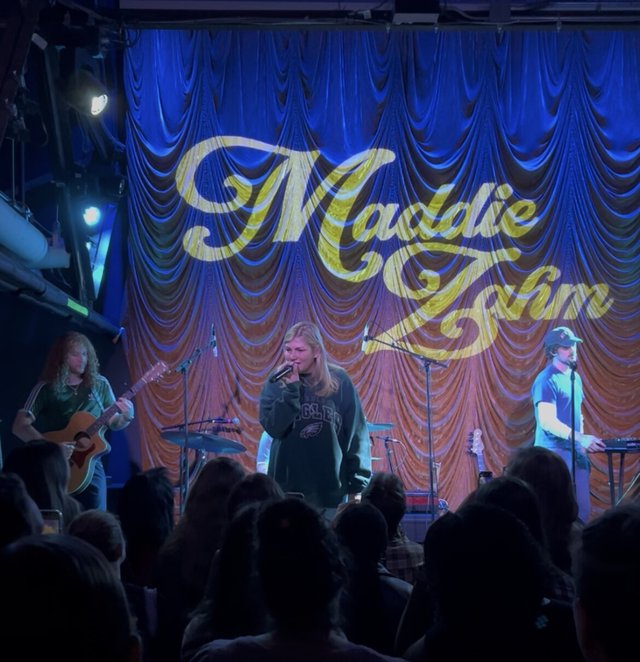 Singer with a microphone on a small stage, flanked by a guitarist and a keyboardist, in front of a blue curtain lit with Maddie Zahm's name