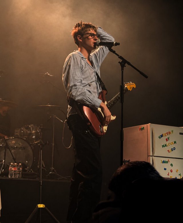 Malcolm Todd in glasses and a light blue shirt singing and playing guitar on a smoky stage