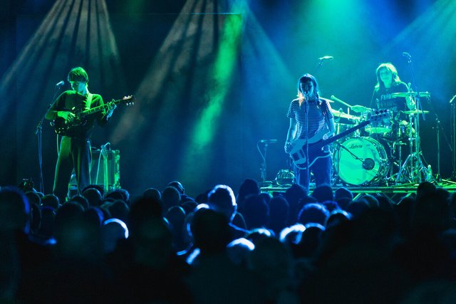 Guitarist, bassist and drummer of a band playing on a stage lit in green and blue, with a silhouetted crowd in front
