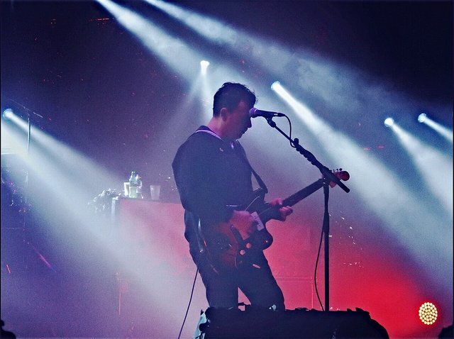 Guitarist silhouetted at a microphone on a smoky stage with beams of white light behind him