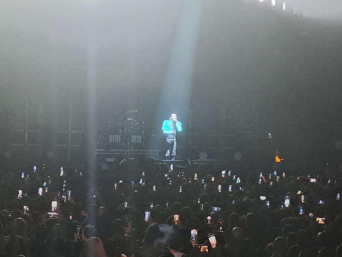Marilyn Manson singing into a microphone under a single white spotlight while the crowd holds up phones