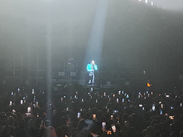 Marilyn Manson singing into a microphone under a single white spotlight while the crowd holds up phones