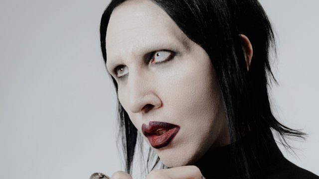 Close-up portrait of Marilyn Manson in white face makeup, dark lipstick and pale contact lenses against a gray backdrop
