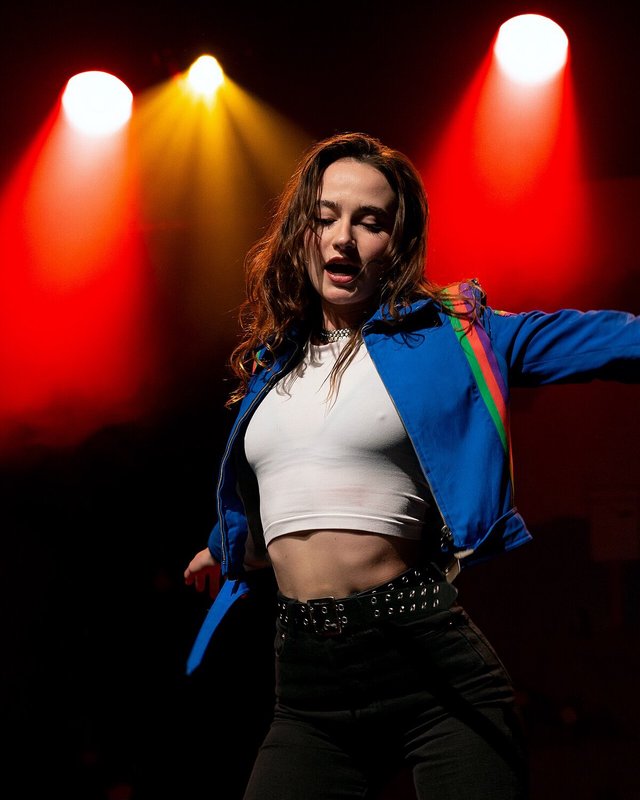 MARIS dancing on stage in a blue jacket under red and amber stage lights