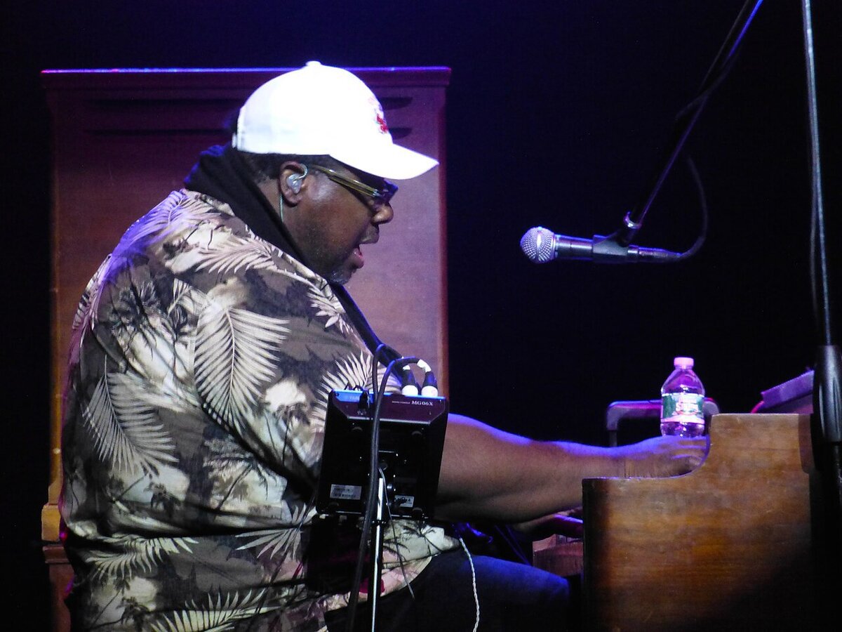 Organist in a white cap and palm-print shirt playing a Hammond organ on a dark stage beside a microphone