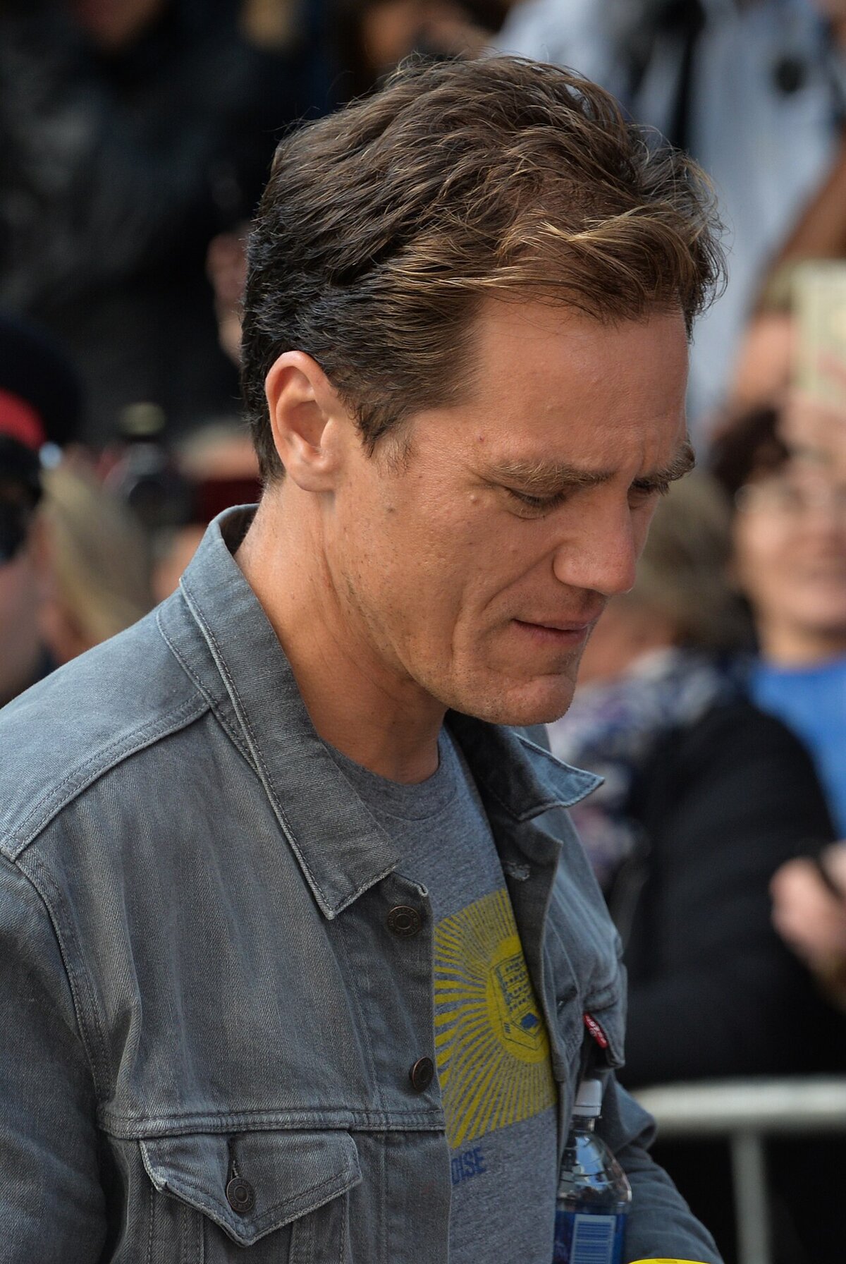 Close-up portrait of Michael Shannon in a gray denim jacket, looking down, with a crowd behind him