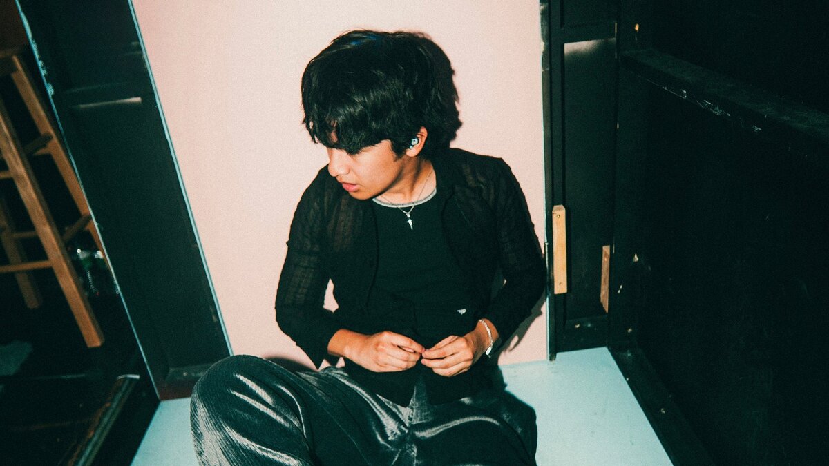 Mico seated on the floor in a black cardigan and silver necklace, looking down and to the side