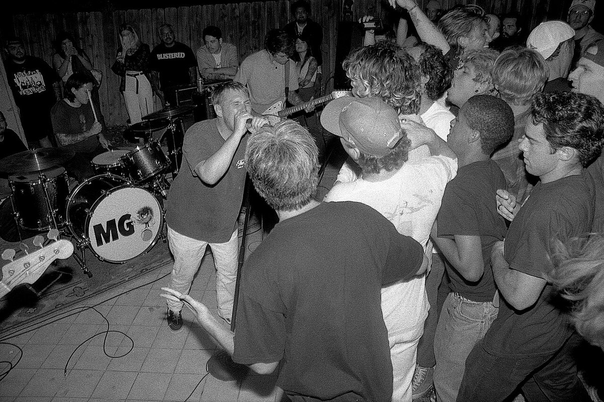 Black-and-white photo of a singer surrounded by a packed crowd beside a drum kit marked MG