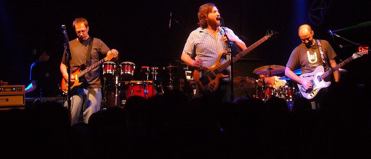 Three members of a jam band playing guitar and bass on a dim stage in front of a silhouetted crowd