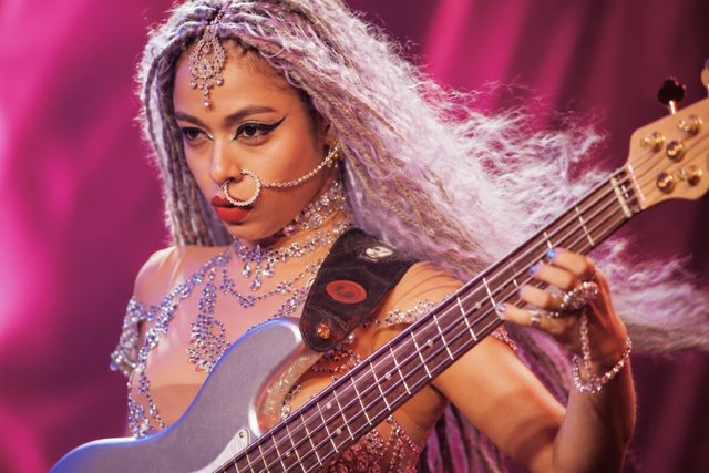 Bassist with long braided hair and jeweled face ornaments plucking a five-string bass under magenta stage light