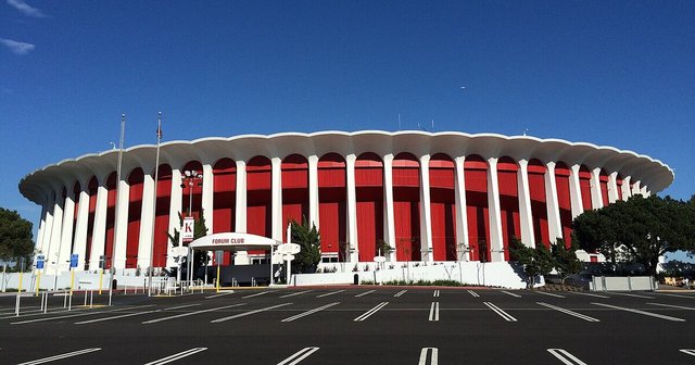 The round, white-columned Kia Forum arena in Inglewood under a clear blue sky, with the Forum Club entrance canopy in front
