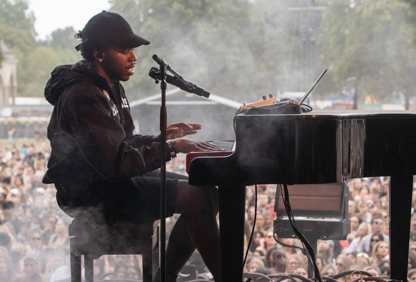 Musician in a black cap and hoodie singing into a microphone while playing a piano on an outdoor stage in front of a crowd