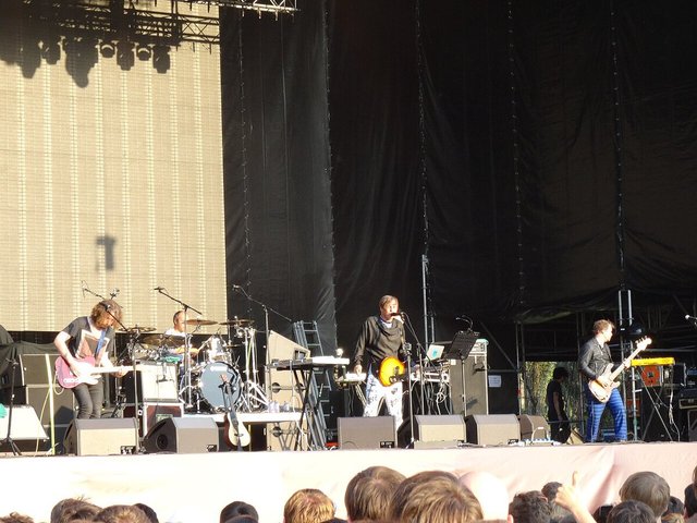 Band playing on a large outdoor stage with a singer at the center microphone, guitarists on either side and a drummer behind, seen over the heads of the crowd
