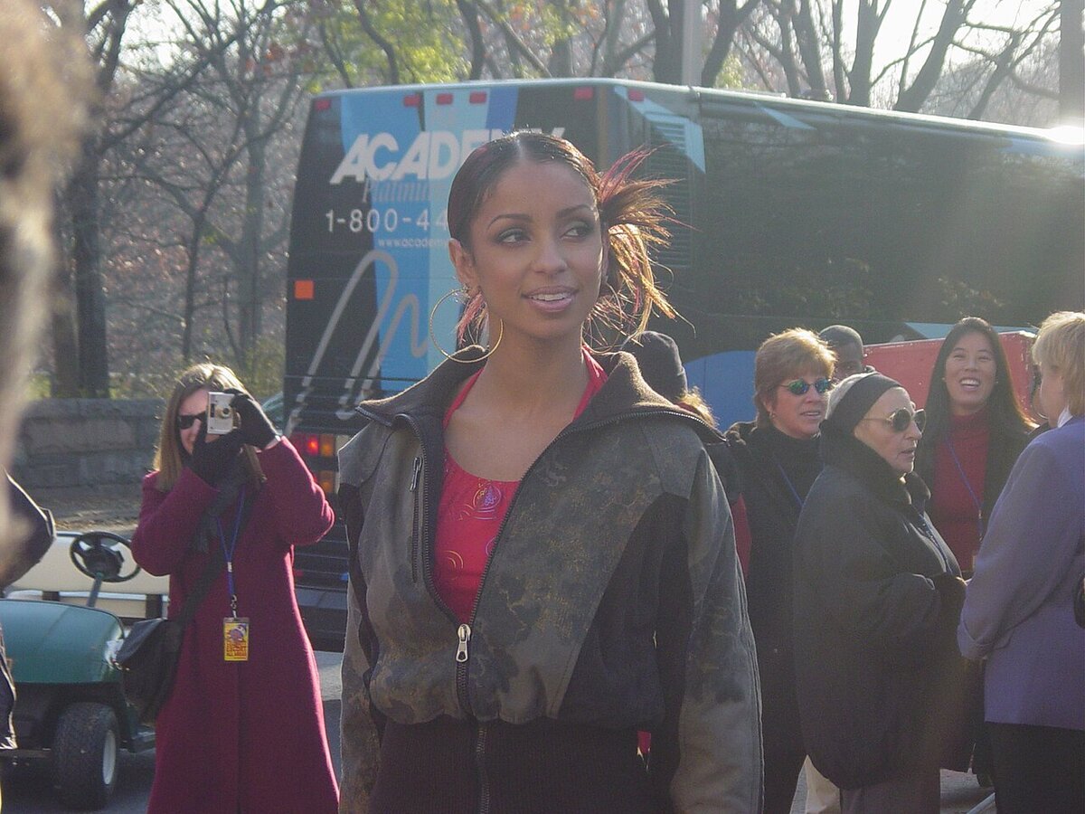 Mýa smiling and looking to the side outdoors, wearing a camouflage-pattern jacket over a red top