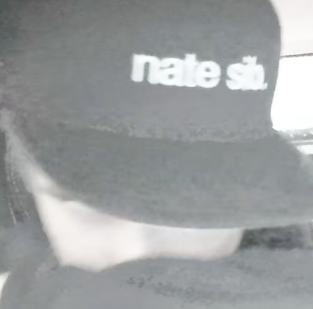 Grainy black-and-white close-up of Nate Sib wearing a dark cap with "nate sib" printed across the front