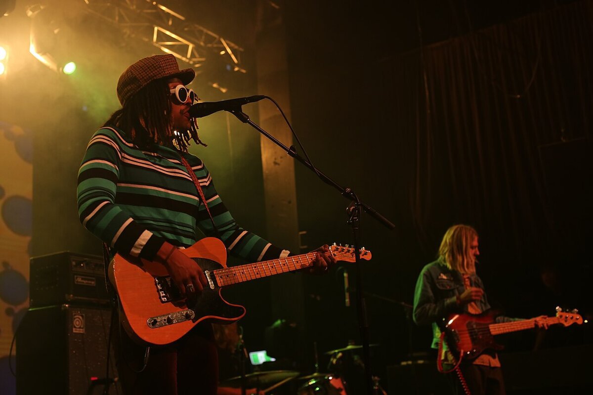 Singer in a patterned sweater and white goggles playing guitar at a microphone while a bassist plays behind, under hazy stage lights