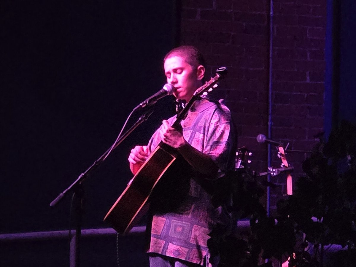 Olive Klug singing into a microphone and playing acoustic guitar on a dimly lit stage with a brick wall behind