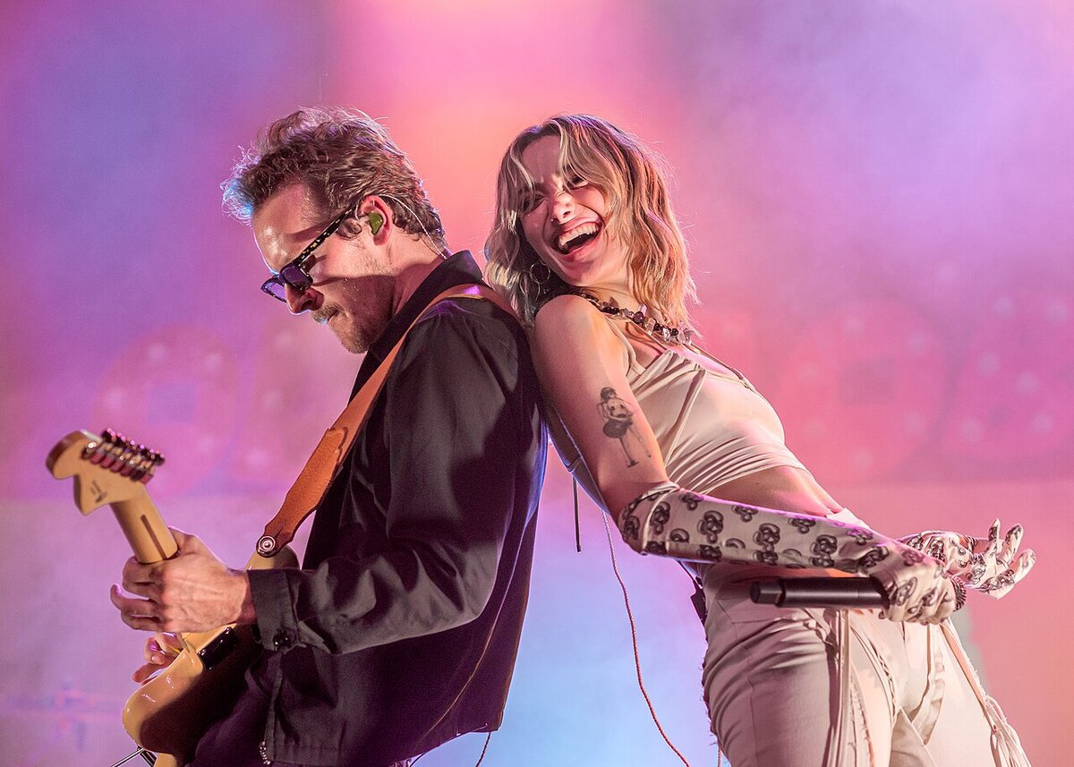 Olivia O'Brien laughing on stage beside a guitarist under pink and purple stage lights