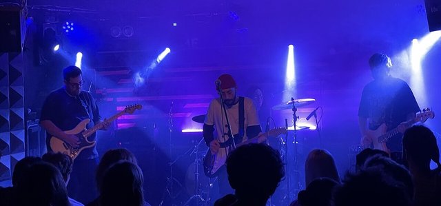 Three members of a band playing guitars and bass under blue stage lights, with the backs of crowd members' heads in the foreground