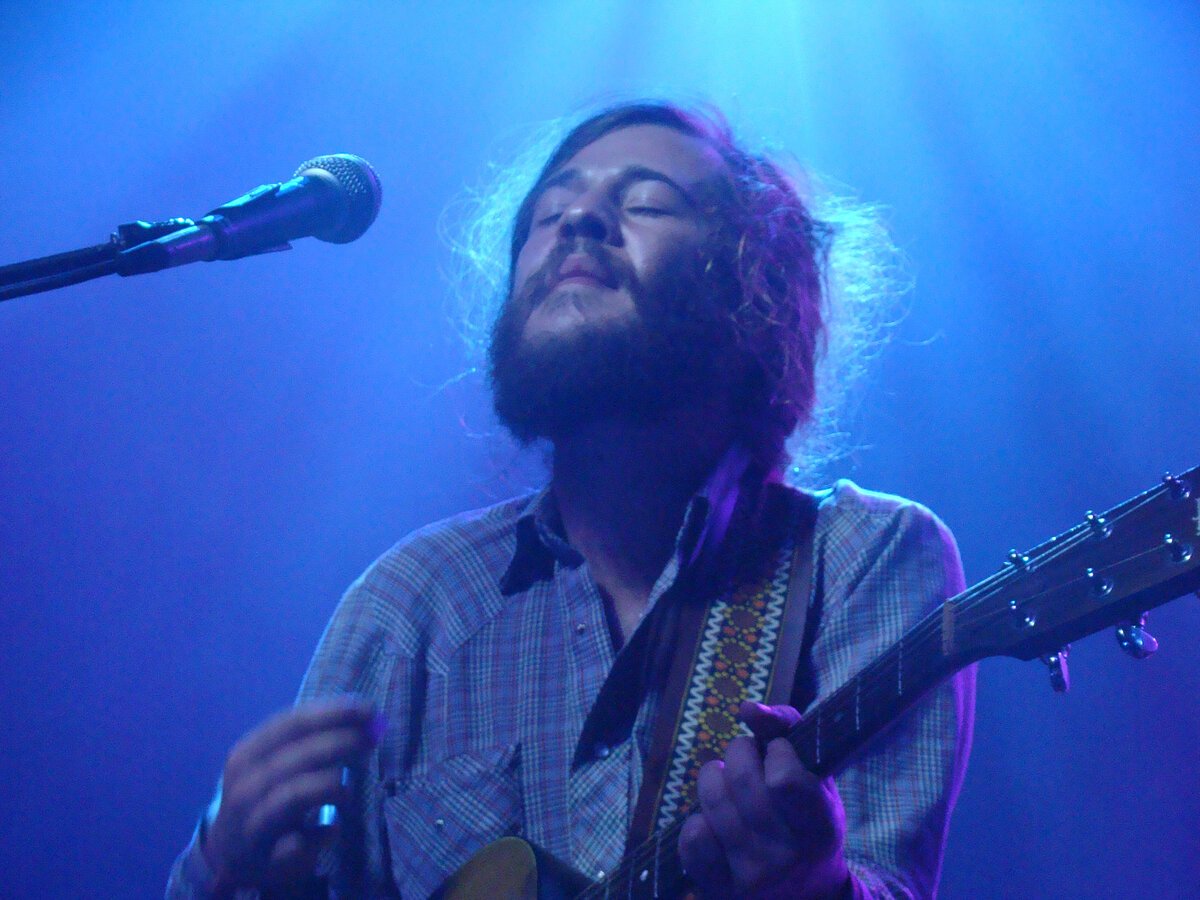 Bearded guitarist singing with eyes closed beside a microphone under blue stage light