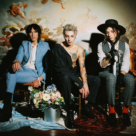 Three members of Palaye Royale posed on chairs in front of a painted ceiling mural