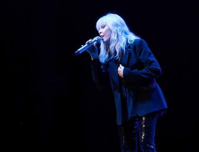 Pat Benatar singing into a handheld microphone on a dark stage, wearing a black blazer and sequined pants under blue light