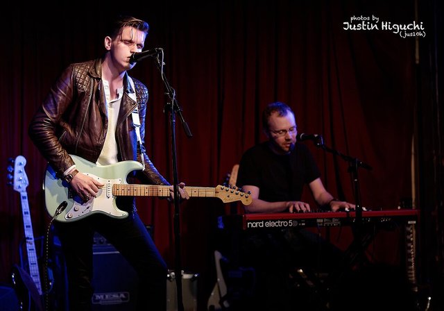Patrick Droney playing a pale green electric guitar on a small stage, with a keyboardist behind him